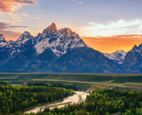Wide view of the Grand Teton mountain range at sunset, with a winding river and forested valley in the foreground.