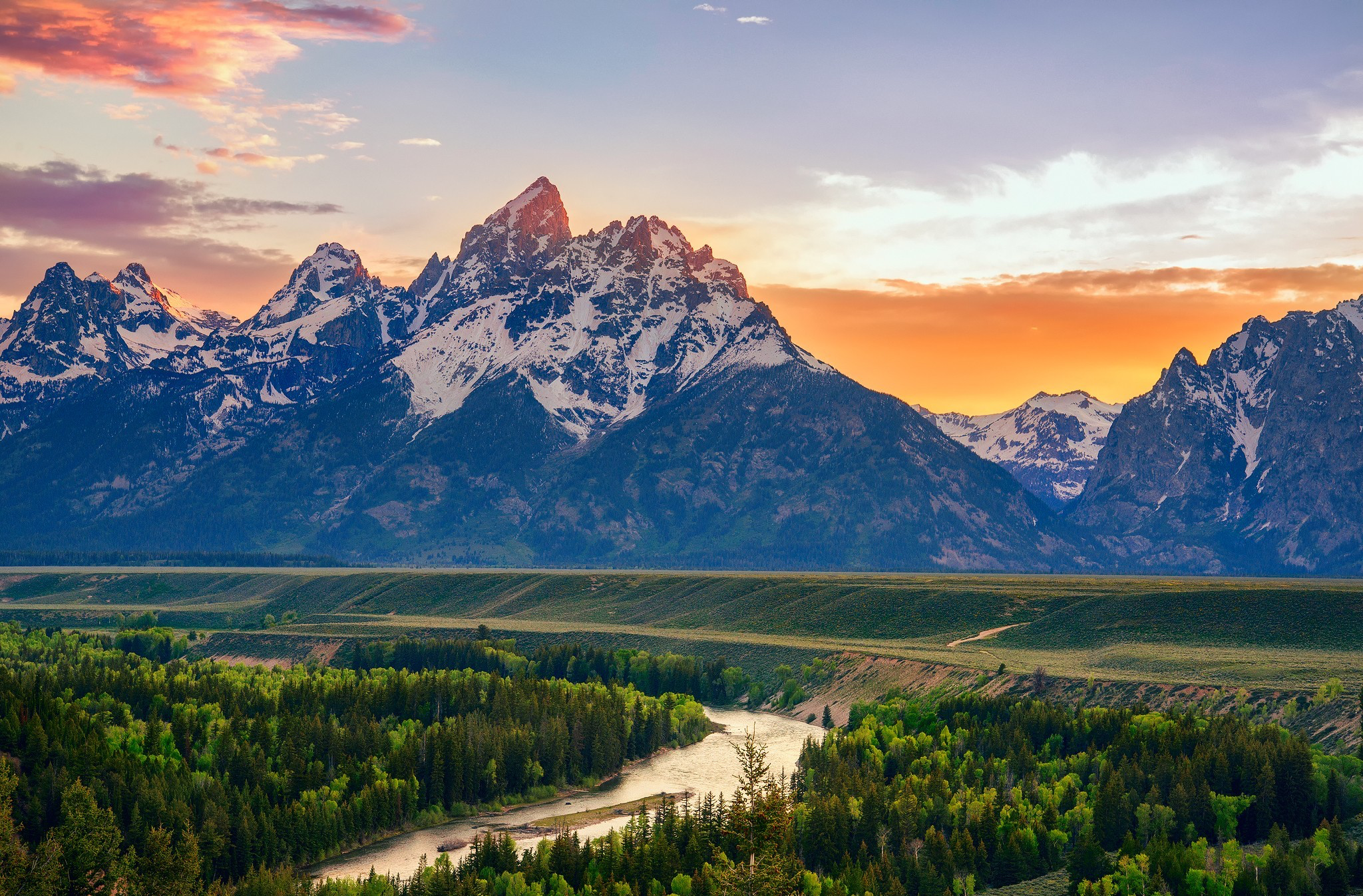 Wide view of the Grand Teton mountain range at sunset, with a winding river and forested valley in the foreground.